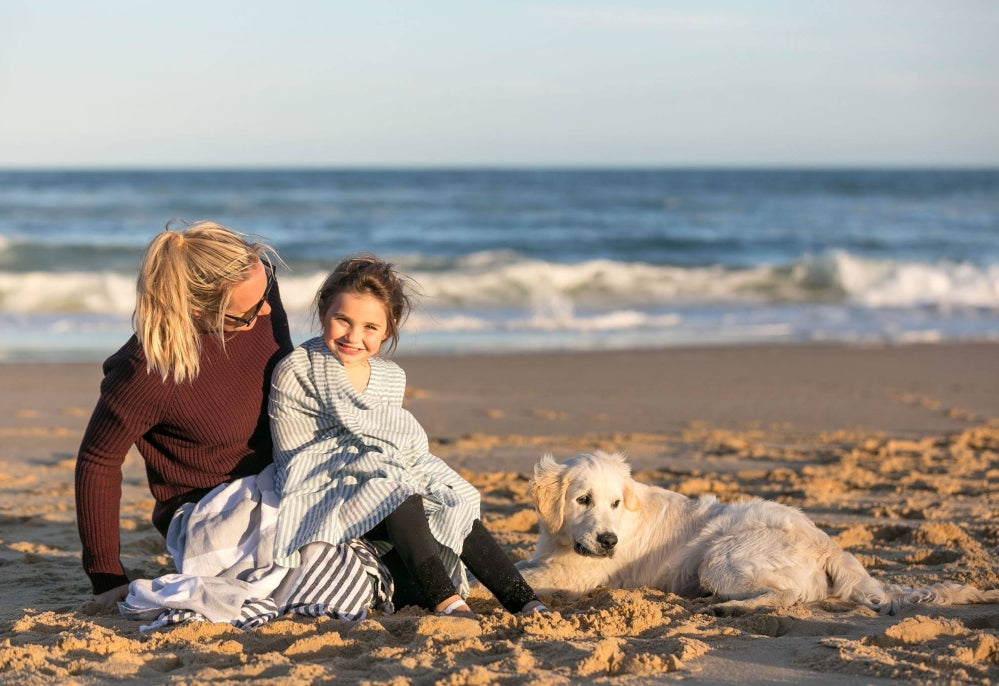 A parent and child sit in the sun on a beach, wrapped in sweaters and a Turkish towel, smiling while a golden lab dog sits beside them