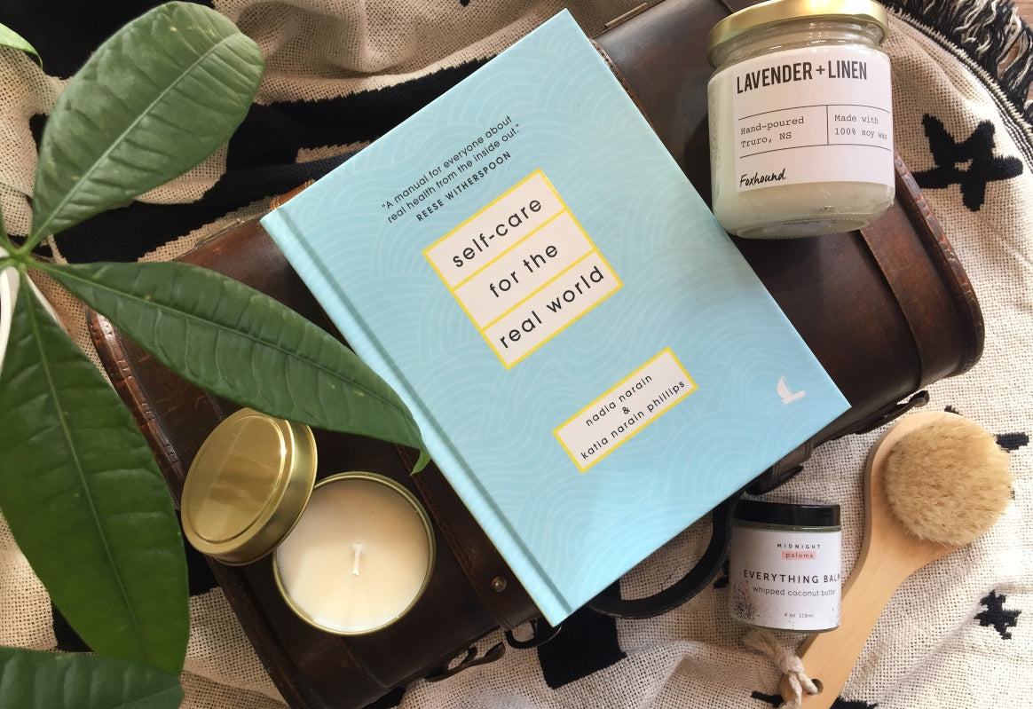 Aqua blue and yellow Self-Care for the Real World book sits between two candles on top of an ivory and navy blanket with green plant leaves hanging over the corner